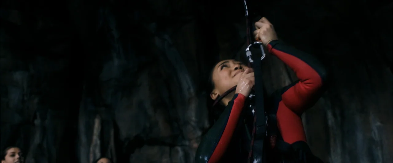 Anaconda (2024) movie still: medium — A dark, cavernous space with wet rock walls and deep shadows. A young woman climbs a rope while wearing a…