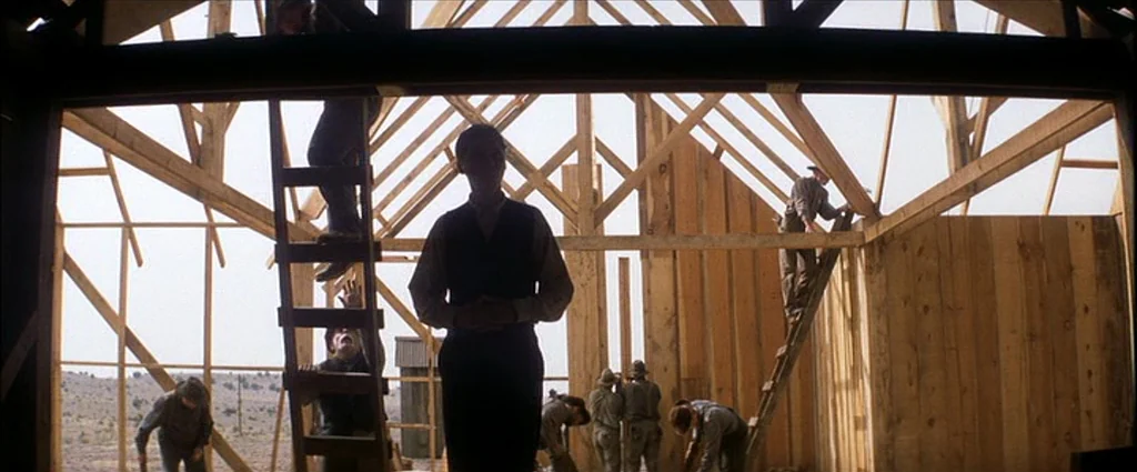 There Will Be Blood (2007) movie still: wide — An unfinished wooden barn structure reveals an open framework against a bright, hazy sky. Several…