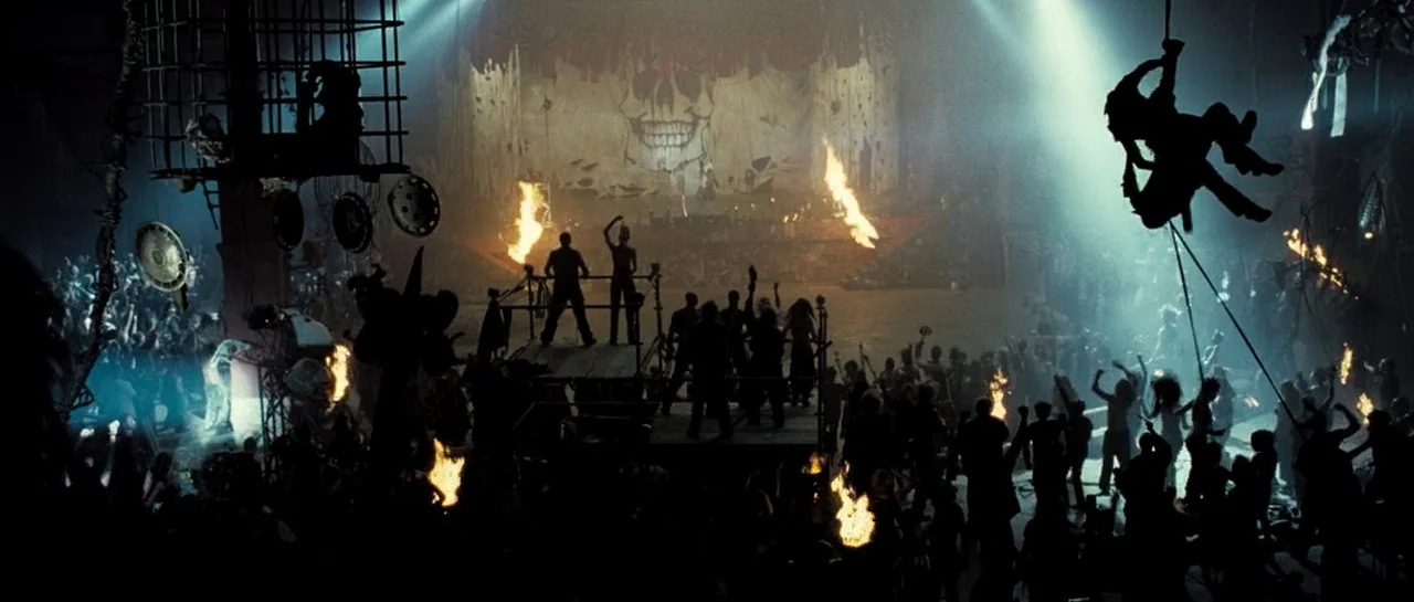 Doomsday (2008) movie still: wide — An industrial hall contains a large central dance floor filled with a dense crowd. A painted skull backdrop…