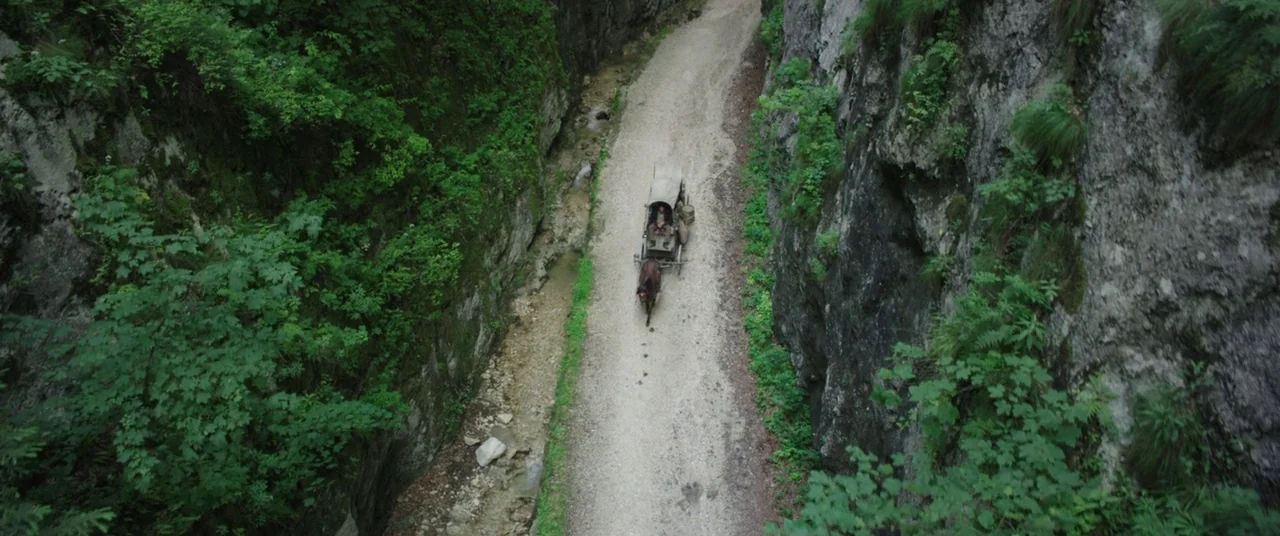 The Nun (2018) movie still: wide — A narrow dirt road runs between two high, steep, plant-covered rock walls. A horse pulls a small, covered wooden…