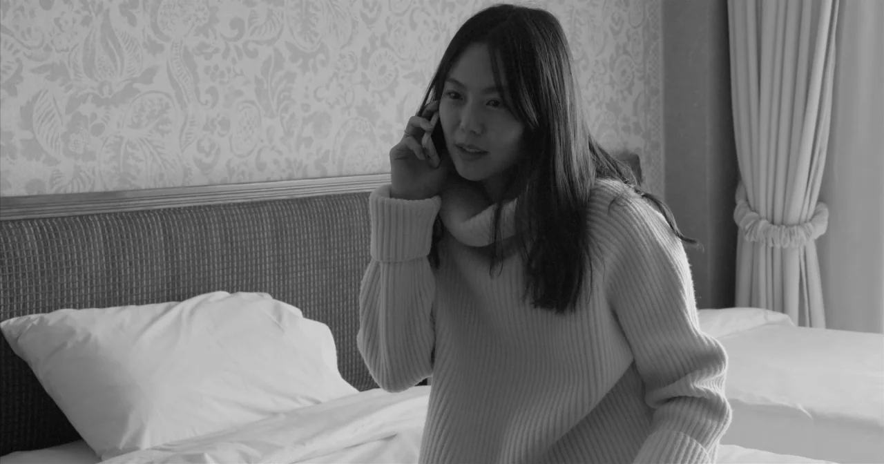 Hotel By The River movie still: medium — A hotel room features a bed with white linens and patterned wallpaper. A young woman sits on the edge of…