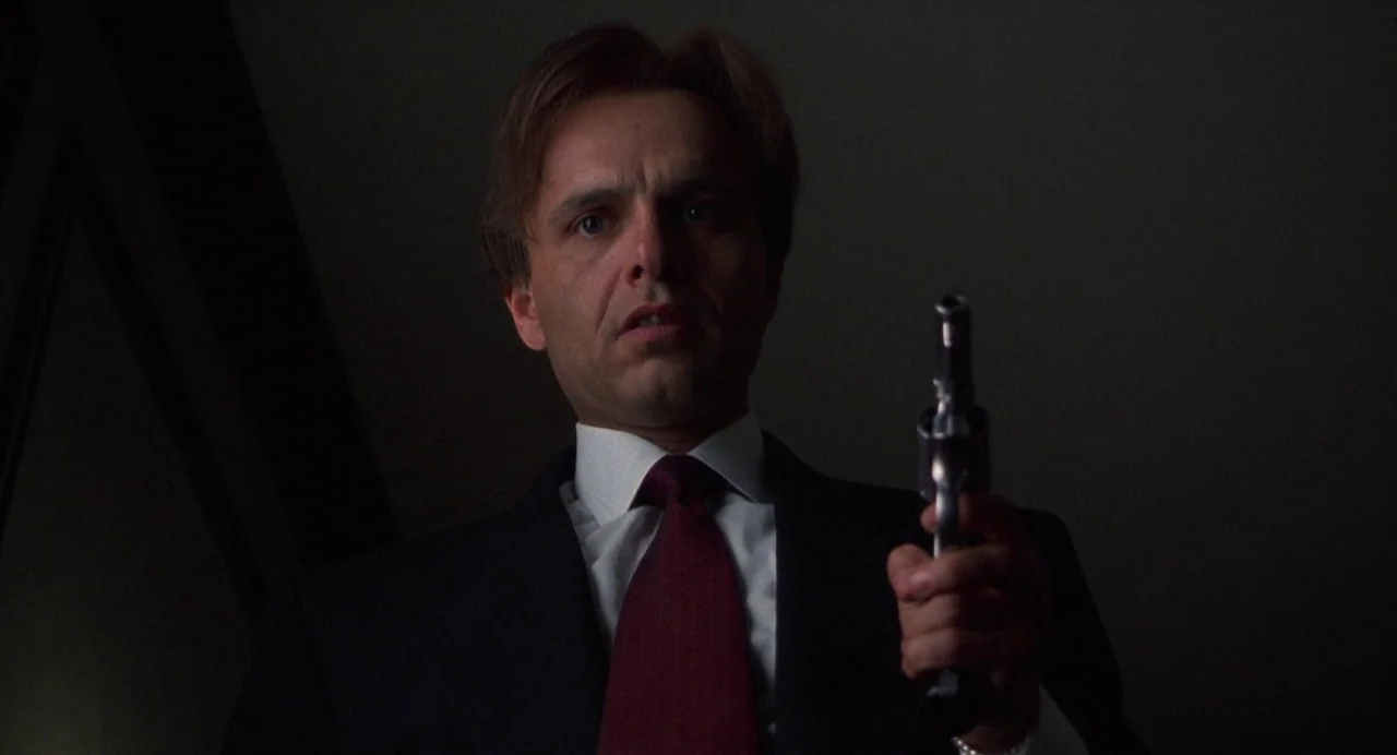 Bound (1996) movie still: medium — A dark, dimly lit interior space with shadows obscuring the background. A man in a suit points a small handgun…