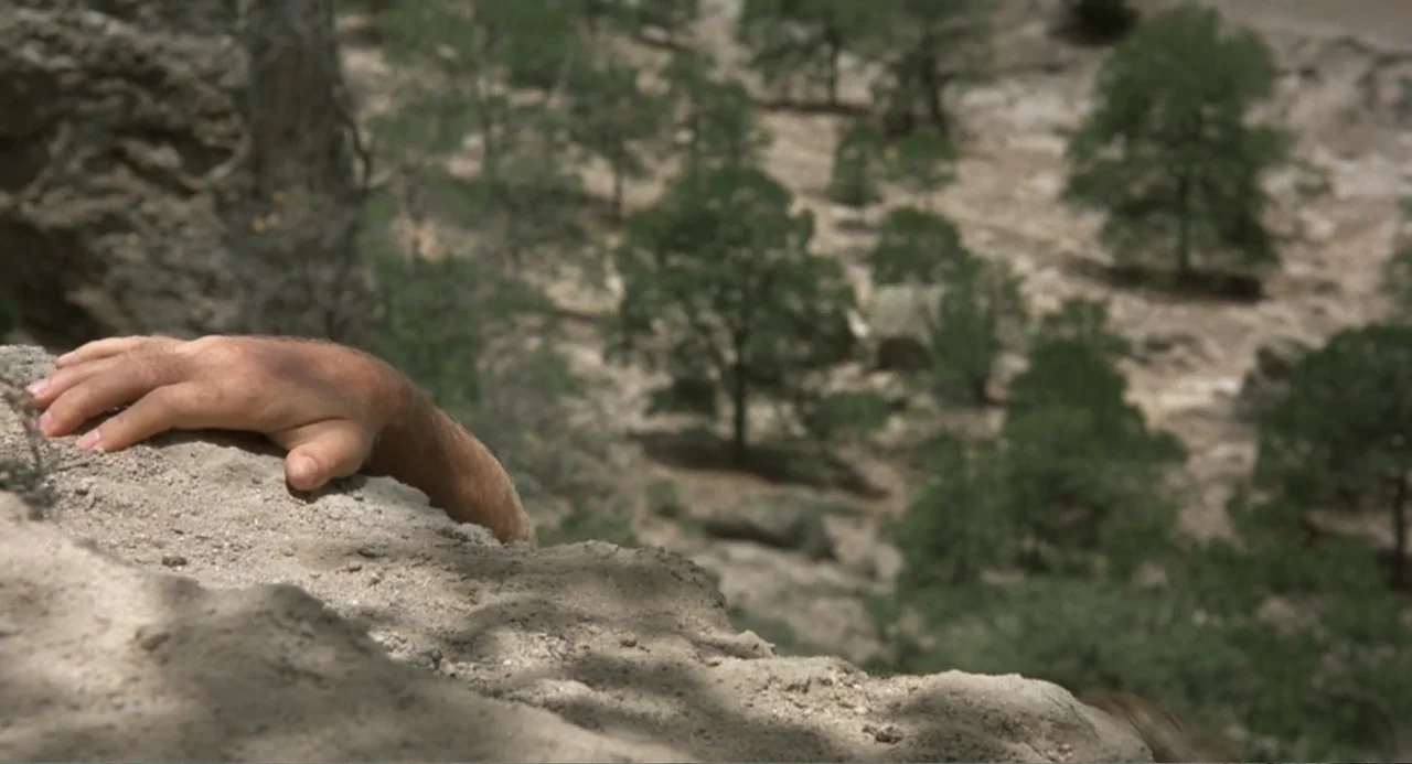 Firewalker (1986) movie still: closeup — A rugged cliff edge overlooks a downward slope filled with scattered pine trees. A human hand grasps the…