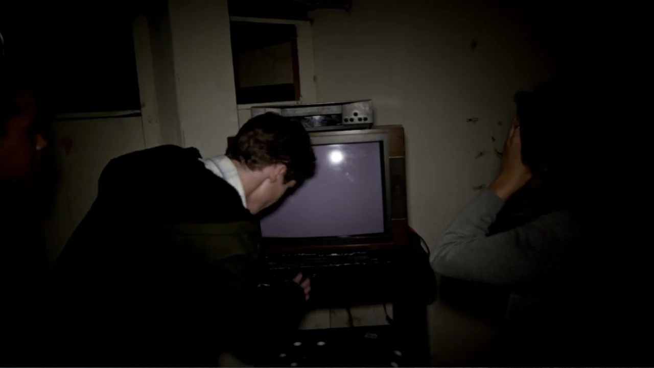 The Gallows (2015) movie still: medium — A dark, unfinished basement with bare walls contains a vintage television and VCR unit. Two individuals…