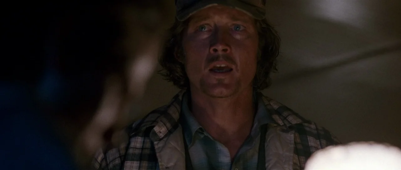 Fire In The Sky (1993) movie still: closeup — A dimly lit interior space with shadows obscuring the background. A man looks toward the left of the…