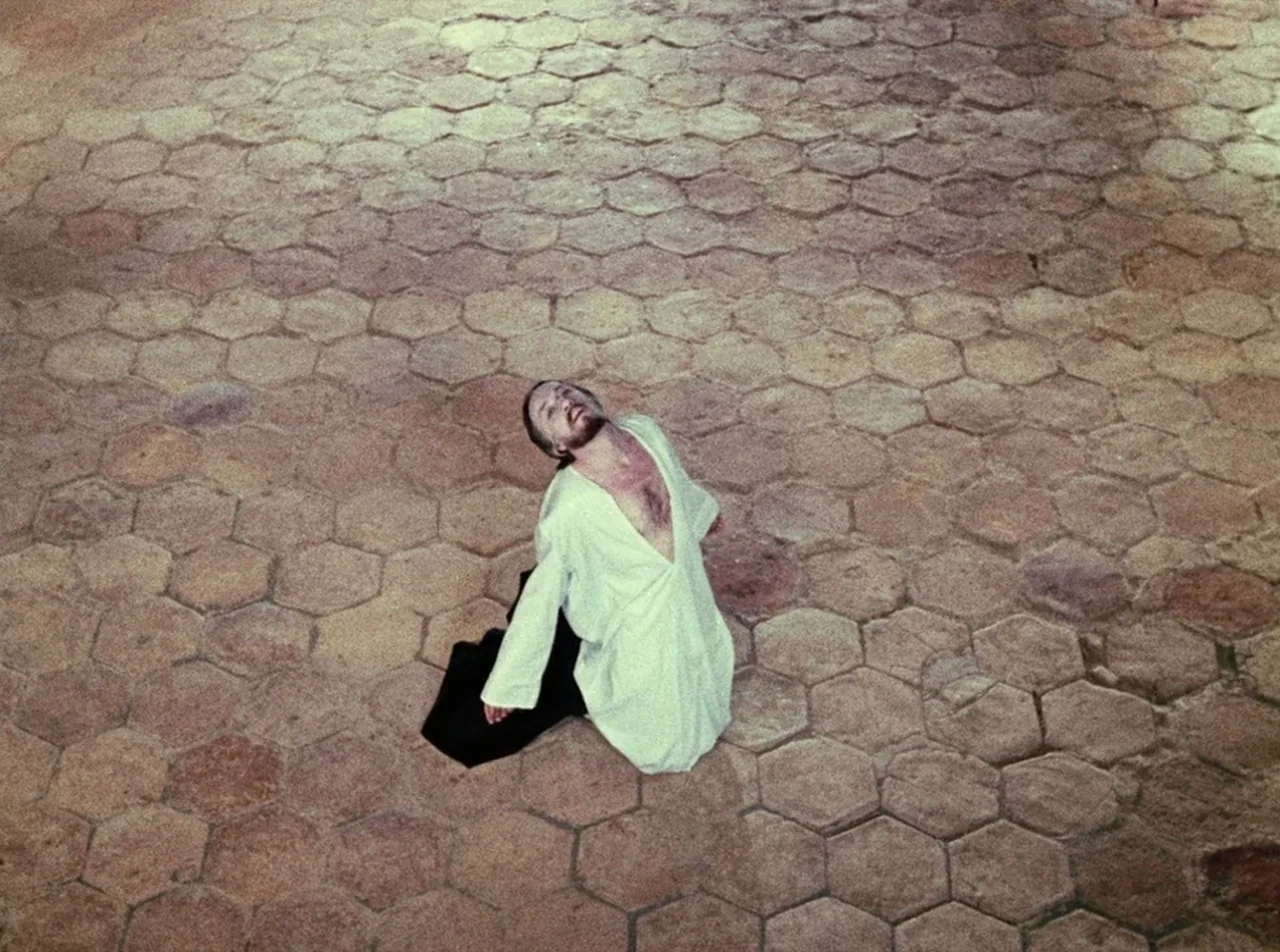 The Colour of Pomegranates (1969) movie still: wide — A man kneels on a vast expanse of light brown hexagonal stone paving. He tilts his head…