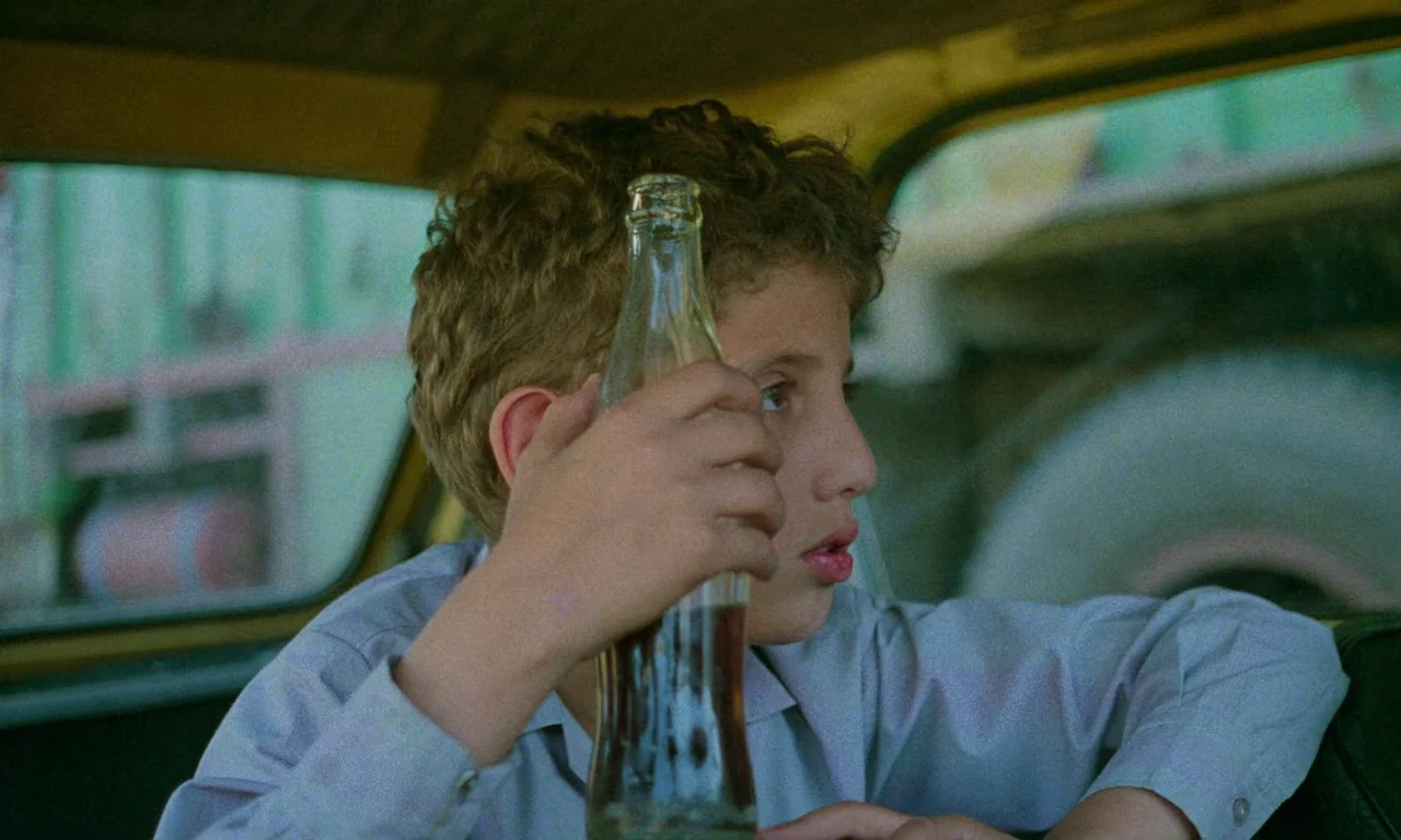 Life, and Nothing More (1992) movie still: medium — Inside a moving vehicle, a young boy with curly hair sits in the passenger seat. He holds a…