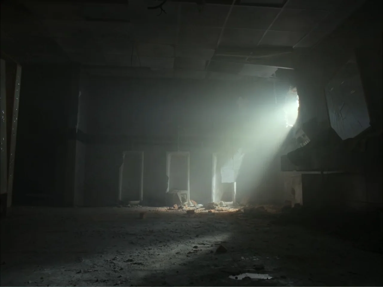 November (2025) movie still: establishing — An abandoned, crumbling restroom features damaged wall partitions and debris scattered across the floor.…