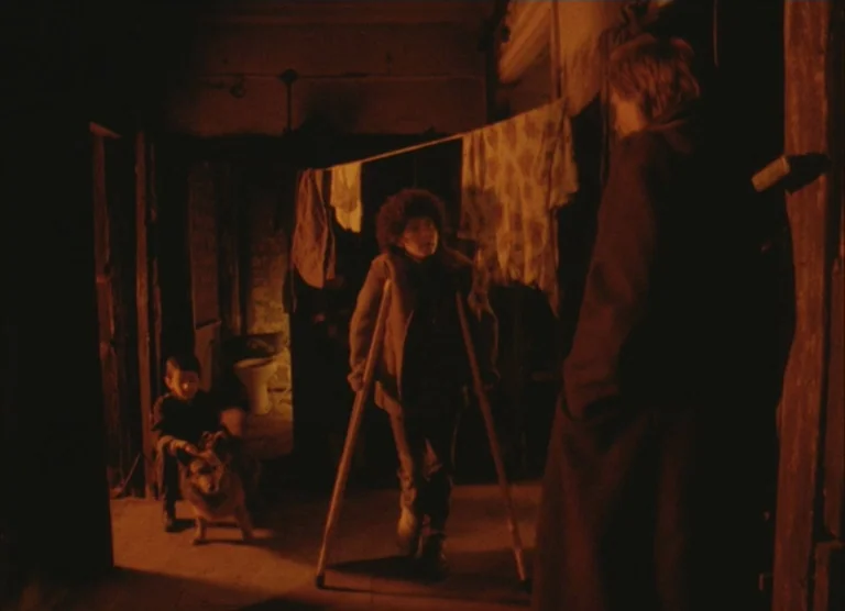 Russian Symphony (1994) movie still: medium — A dark, cramped, and rustic interior space features laundry hanging on a line. A person on crutches…