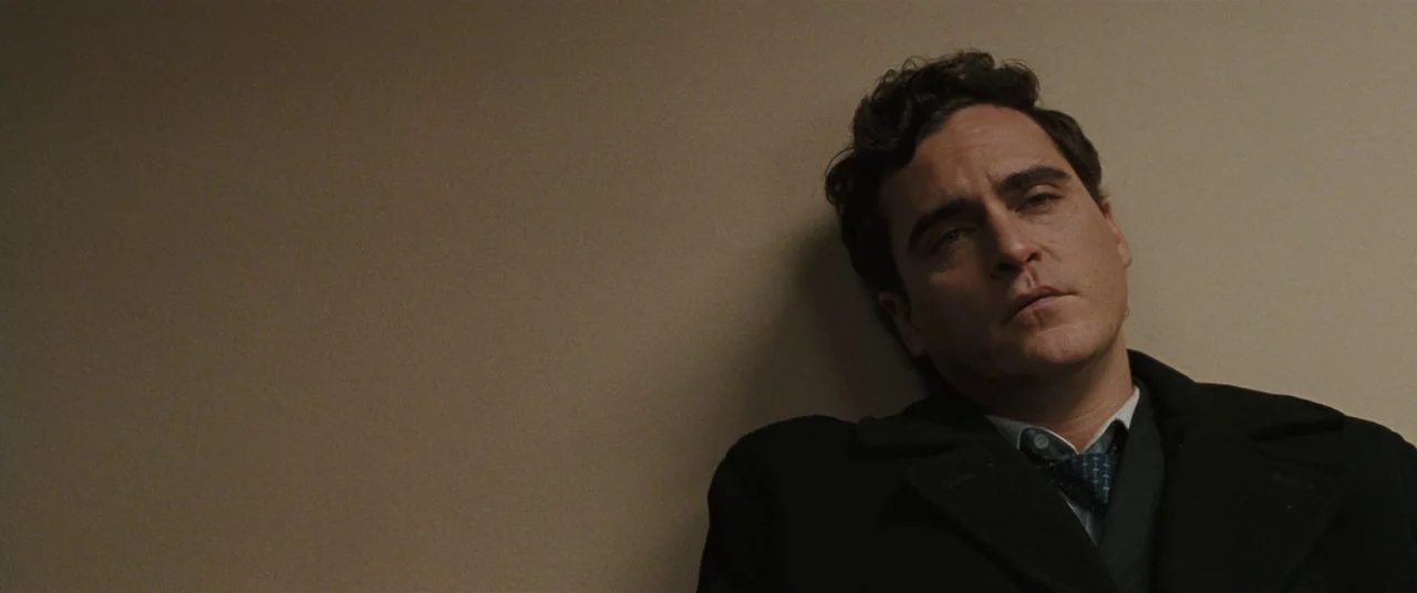 Two Lovers (2008) movie still: medium — A man stands against a plain, off-white wall surface in a minimally lit space. He leans his body back…