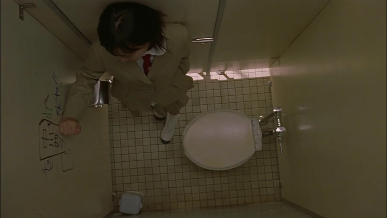 Battle Royale (2000) movie still: fullbody — A cramped tiled public restroom stall featuring beige partition walls with black marker graffiti. A…