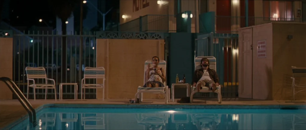 Away We Go (2009) movie still: wide — A dark night pool deck at a motel features two lounge chairs side-by-side. A man and woman sit in the chairs…