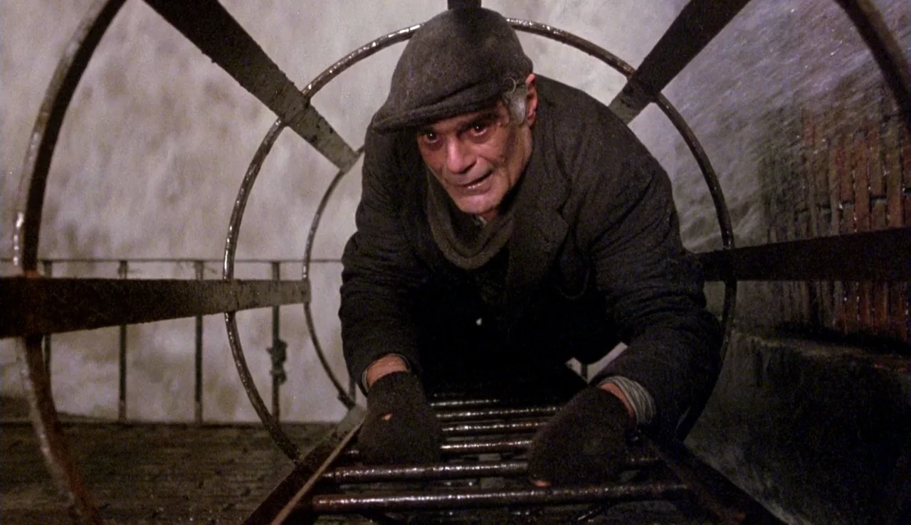 The Rainbow Thief (1990) movie still: medium — A circular metal ladder structure sits within a damp stone or brick tunnel. An elderly man in a cap…