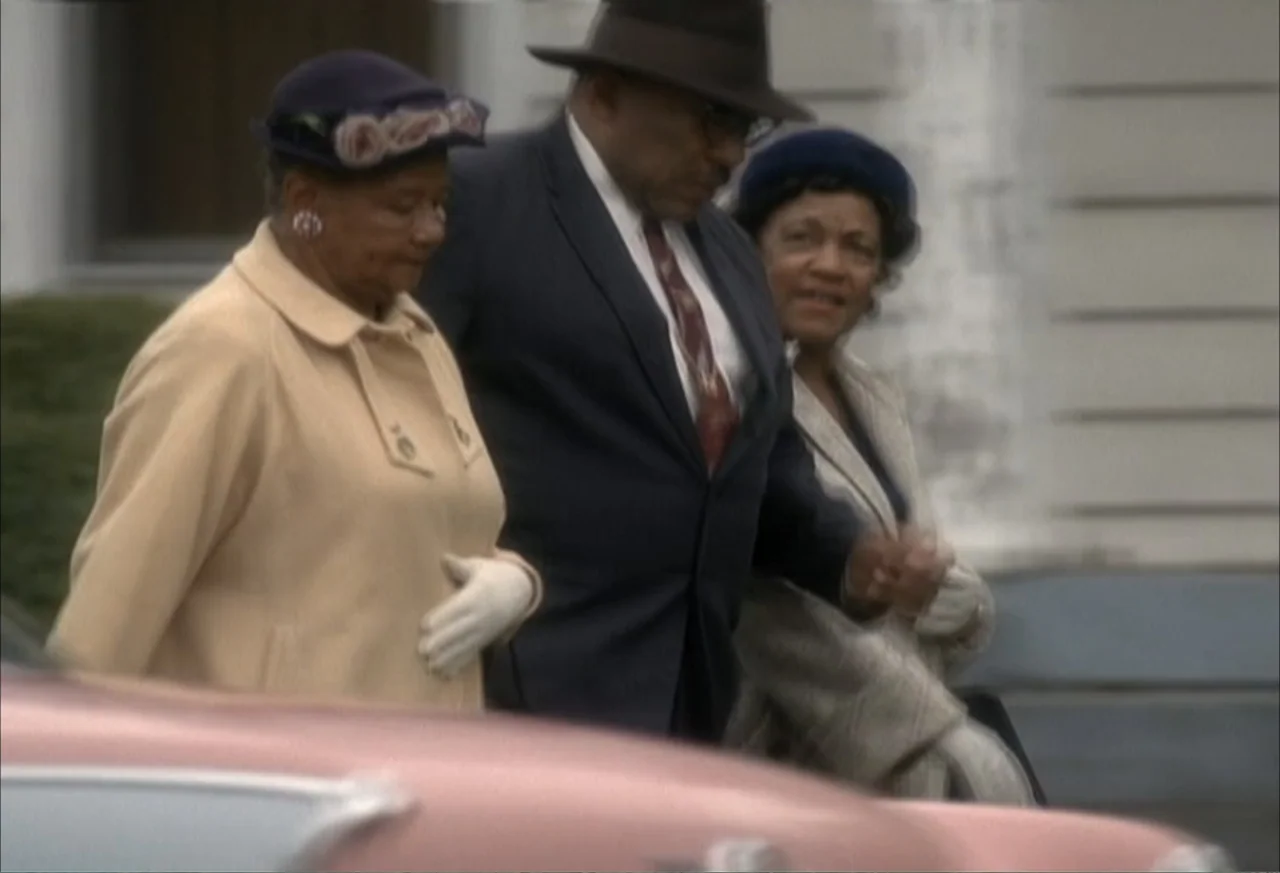 Boycott (2001) movie still: medium — A group of three well-dressed individuals walk past a pink vintage car. They are positioned on a sidewalk in…