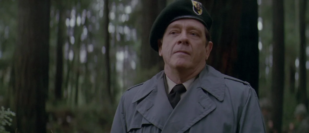 First Blood (1982) movie still: medium — A man in a military beret and gray trench coat stands in a woodland. Thick tree trunks surround him in the…