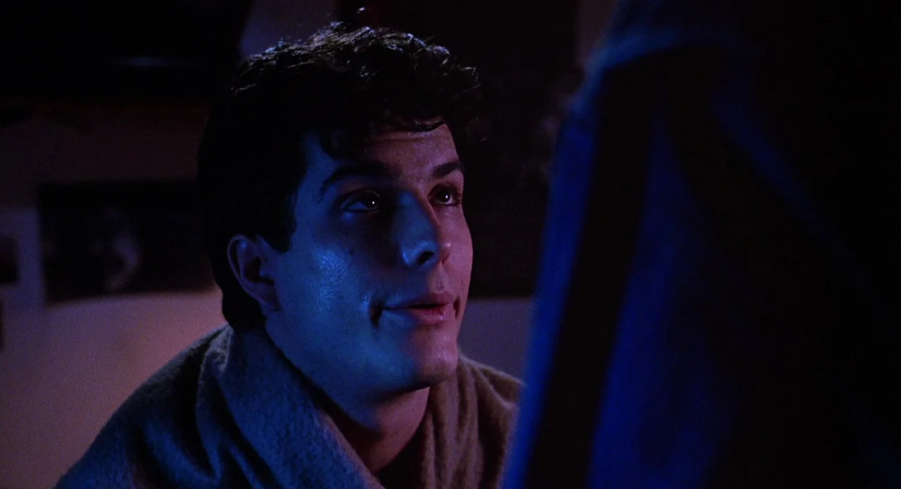 Brain Damage (1988) movie still: closeup — A dimly lit room with heavy shadows and blue light cast on a subject. A young man with dark curly hair…