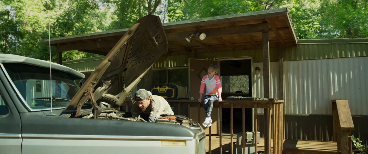 Logan Lucky (2017) movie still: wide — A man repairs the engine of a truck parked in front of a mobile home. A young girl sits on the wooden porch…