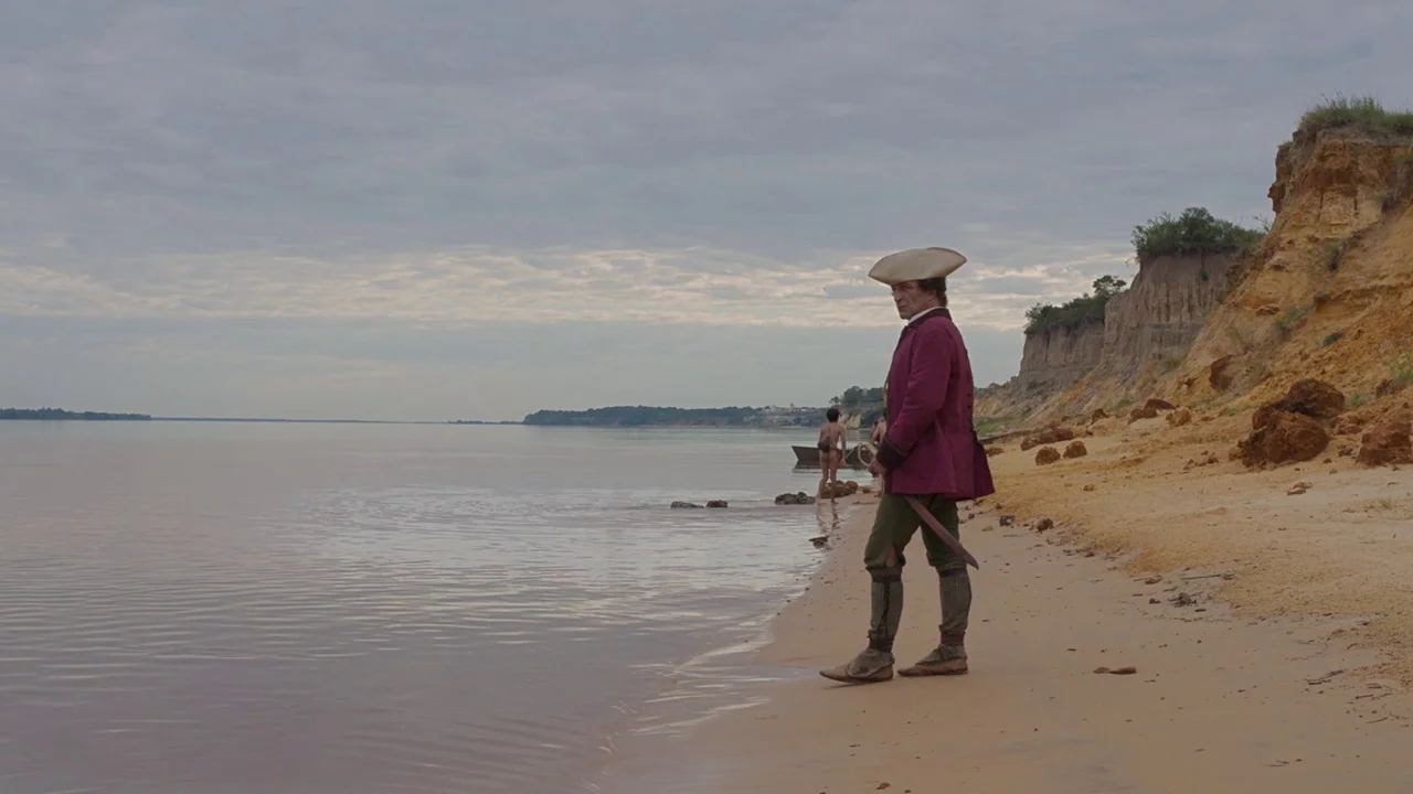 Zama (2017) movie still: fullbody — A sandy beach meets a wide river under a heavily clouded overcast sky. A man in period-appropriate attire stands…