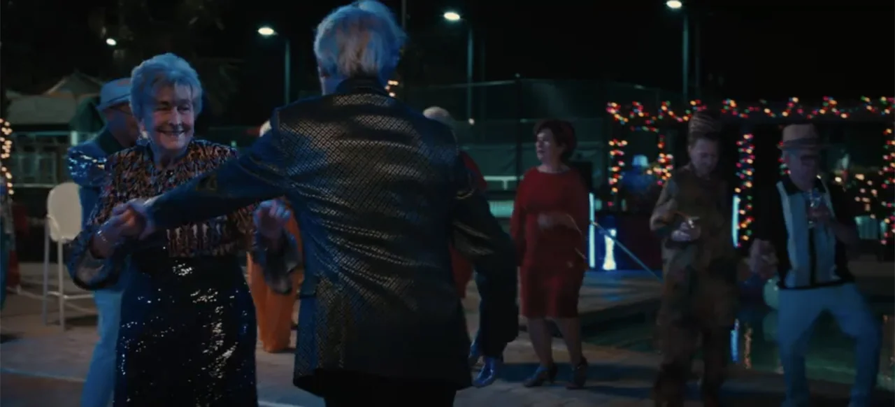 Merv (2025) movie still: medium — An outdoor evening gathering features an elderly couple dancing on a paved poolside area. Festive string lights…