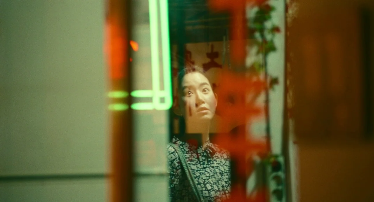 Past Lives (2023) movie still: medium — A view through a glass window showing a woman standing inside a dimly lit area. Red and green lights create…