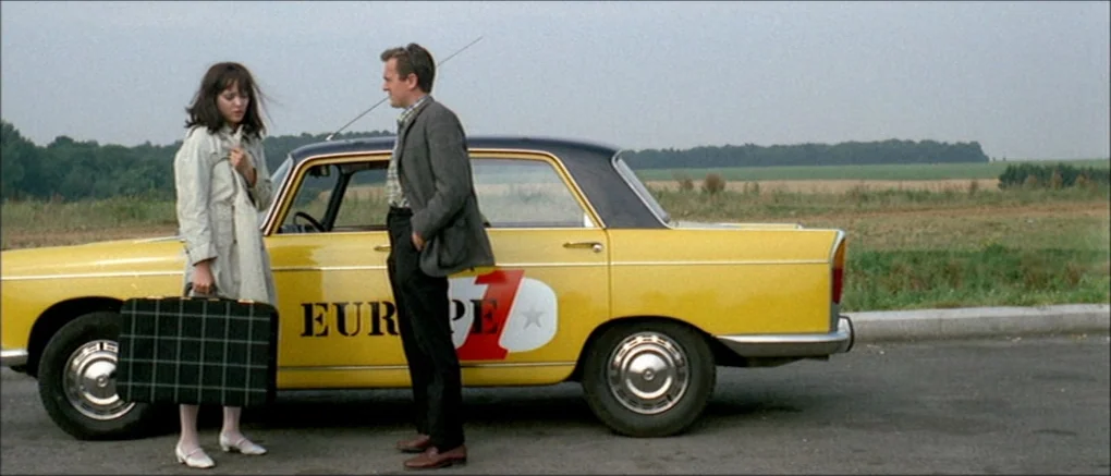 Made in U.S.A. (1966) movie still: fullbody — A man and woman stand beside a yellow sedan parked on a roadside. The car features large black text…