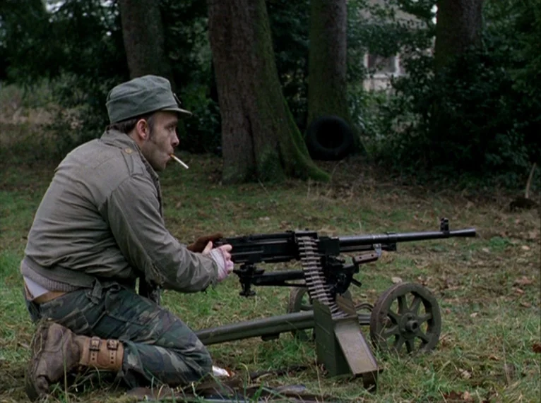 For Ever Mozart (1996) movie still: medium — A man in military-style gear kneels in a grassy area surrounded by mature trees. He holds a belt-fed…
