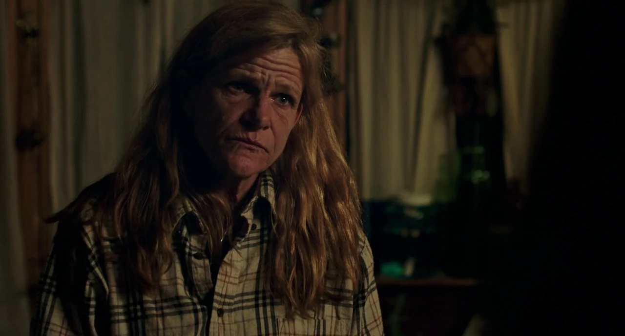 Leave No Trace (2018) movie still: medium — A dimly lit room with white curtains and a partially visible plant on a shelf. A middle-aged woman in a…