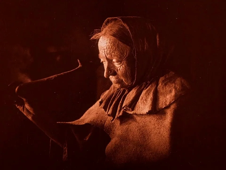 Häxan (1922) movie still: closeup — An elderly woman in rustic clothing sits in a dark, shadow-filled environment. She holds a small, curved object…