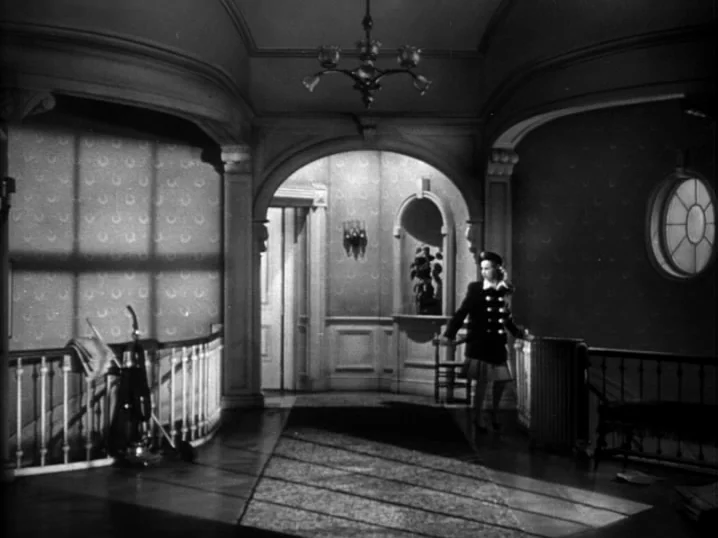 The Seventh Victim (1943) movie still: wide — A spacious home landing features wallpaper patterns and an archway leading to another room. A young…