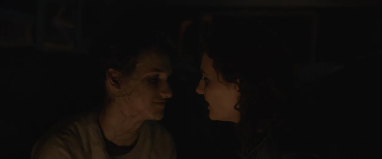 Maggie (2015) movie still: medium — A dark, nondescript interior space with minimal light visibility. Two individuals stand close together in…
