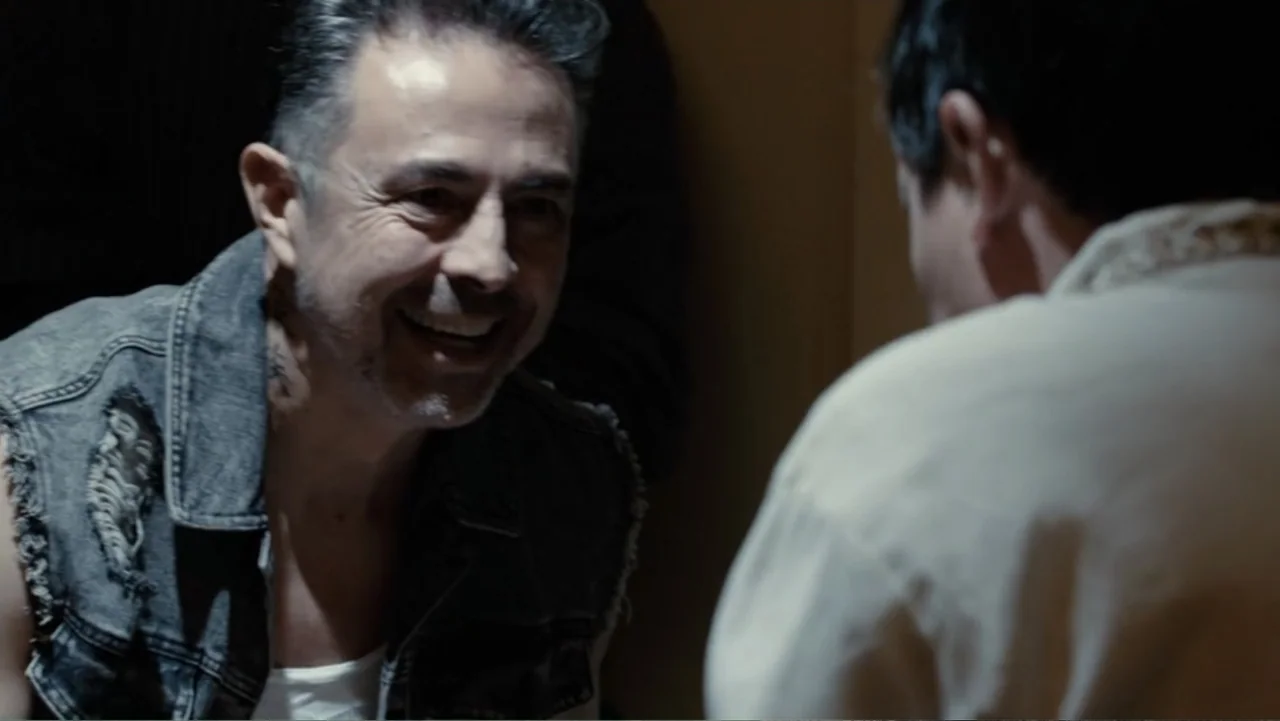 Por Amor al Money (2023) movie still: medium — A dimly lit interior space features two people engaged in a private conversation. The foreground…