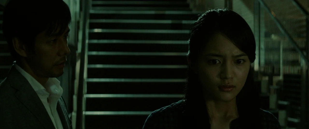 Creepy (2016) movie still: medium — A dark, industrial-style staircase provides the backdrop for the two figures. The man and woman remain…
