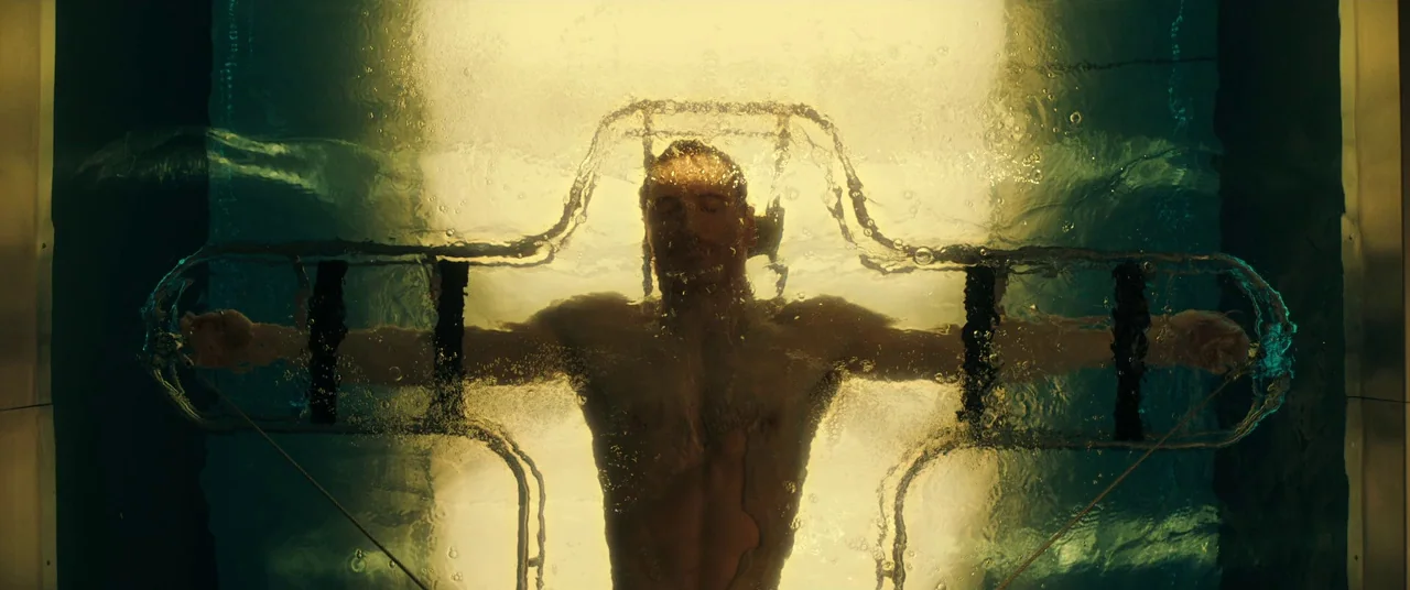 Assassin’s Creed (2016) movie still: medium — A man floats inside a glass-walled tank filled with water and illuminated from behind. He is secured…