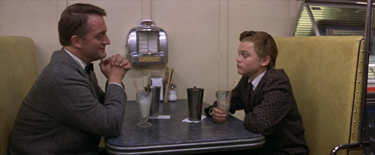 This Boy's Life (1993) movie still: medium — A 1950s style diner booth with yellow vinyl seating and a grey laminate table. An adult man and a young…