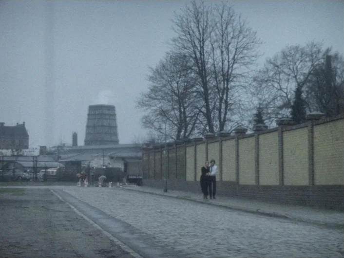 Eika Katappa (1969) movie still: wide — An urban cobblestone street with a long yellow brick wall and bare trees. A man and woman walk alongside the…