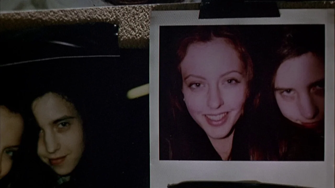 Ginger Snaps (2000) movie still: closeup — A close-up view of two printed photographs pinned to a textured wall. The images show close-up portraits…