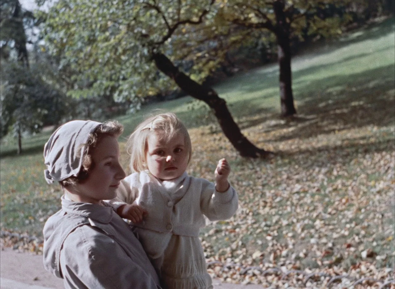 The Red Balloon (1956) movie still: medium — An outdoor park setting with a grassy slope and mature trees in autumn. A young woman carries a blonde…