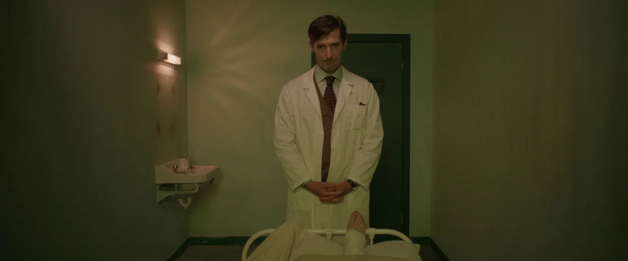Oddity (2024) movie still: medium — A small clinical room has pale green walls and a wall-mounted basin. A doctor in a white lab coat stands behind…