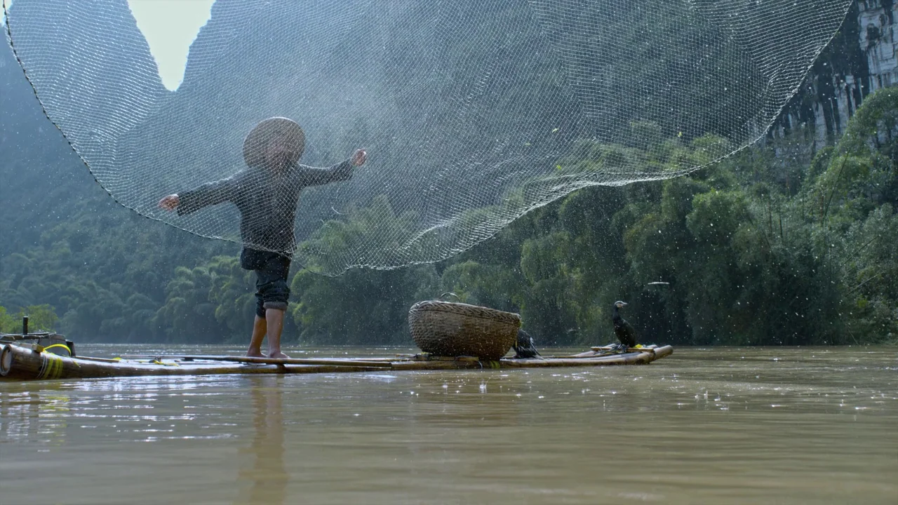 Awaken (2018) movie still: fullbody — A man stands on a bamboo raft on a calm brown river with karst mountains. The fisherman tosses a large…