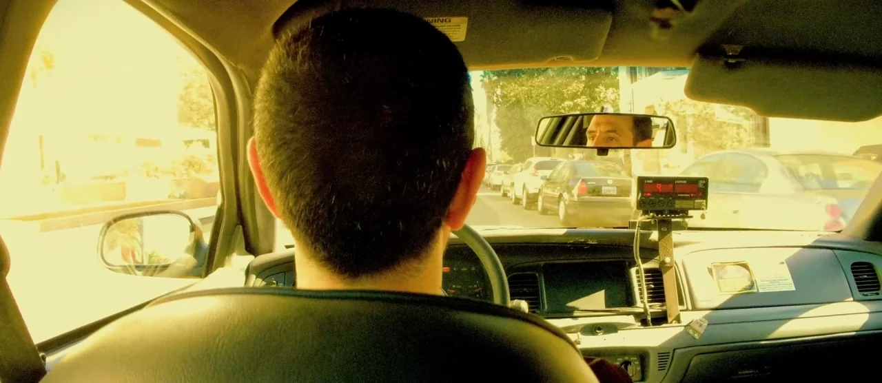 Tangerine (2015) movie still: medium — Inside a taxi, the driver looks ahead while a passenger is visible in the mirror. Outside the windshield, a…
