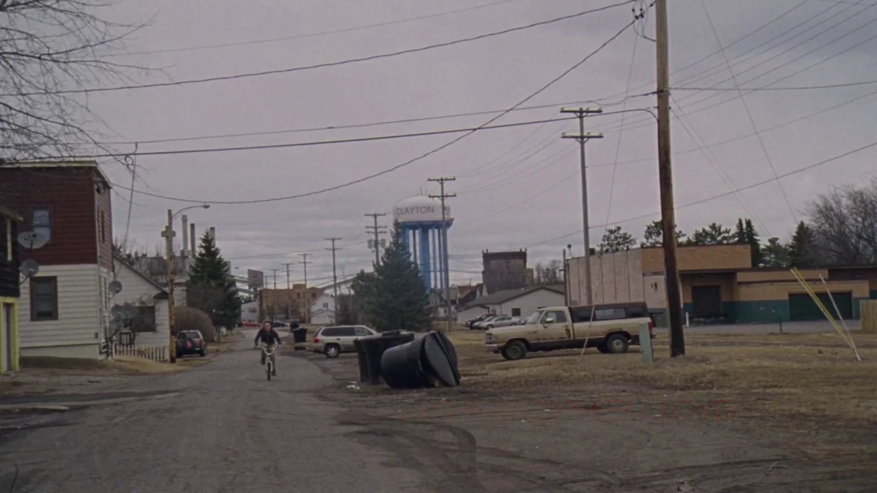 I Am Not a Serial Killer (2016) movie still: wide — A rural street stretches toward a distant water tower labeled Clayton under overcast skies. A…