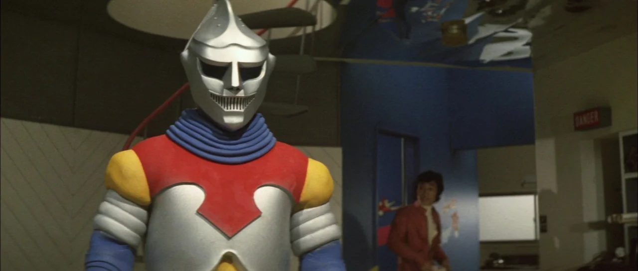 Godzilla Vs Megalon (1973) movie still: medium — A room with blue walls and a white zig-zag wall pattern in the background. A silver-armored robot…