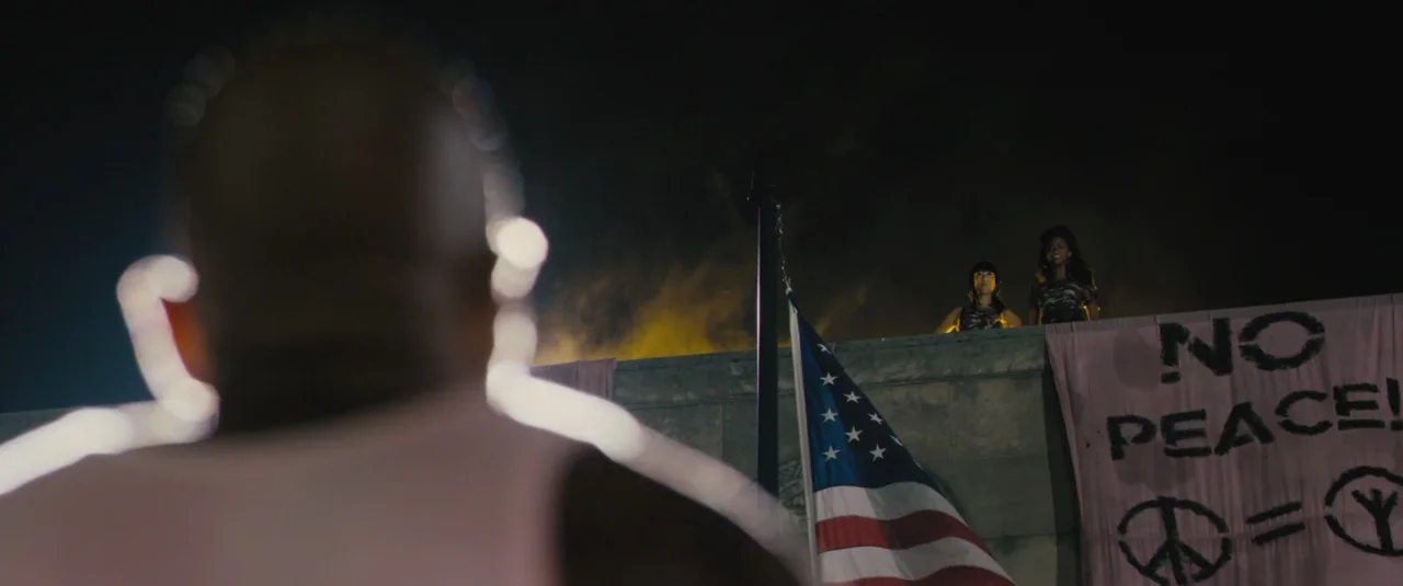 Chi-raq (2015) movie still: wide — A nighttime rooftop with a stone ledge, a flag, and a political banner. Two women stand on the ledge as a man…