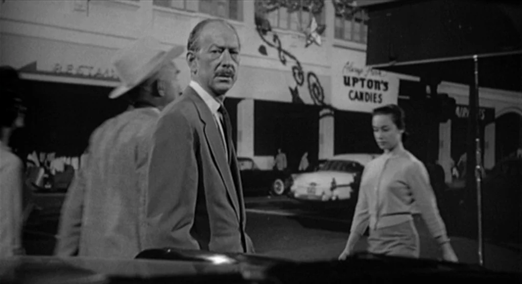 Psycho movie still: medium — A nighttime commercial street with parked 1950s cars and storefront signage. A man stands in the foreground while a…