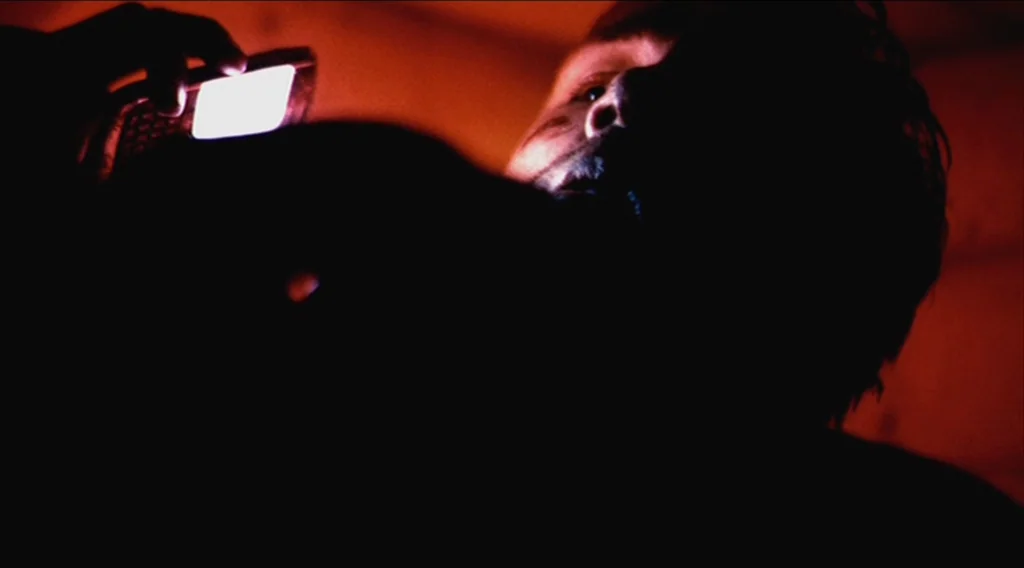 Buried (2010) movie still: closeup — A dark, unidentifiable space is shrouded in near-total shadow. A person holds a bright mobile phone that…