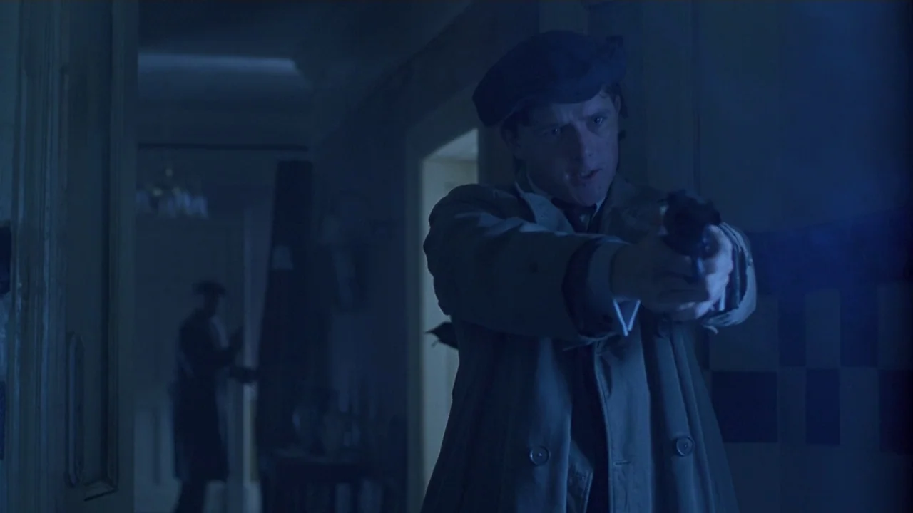 Michael Collins (1996) movie still: medium — A dimly lit narrow hallway at night with dark blue shadows. A man in a trench coat and flat cap holds a…