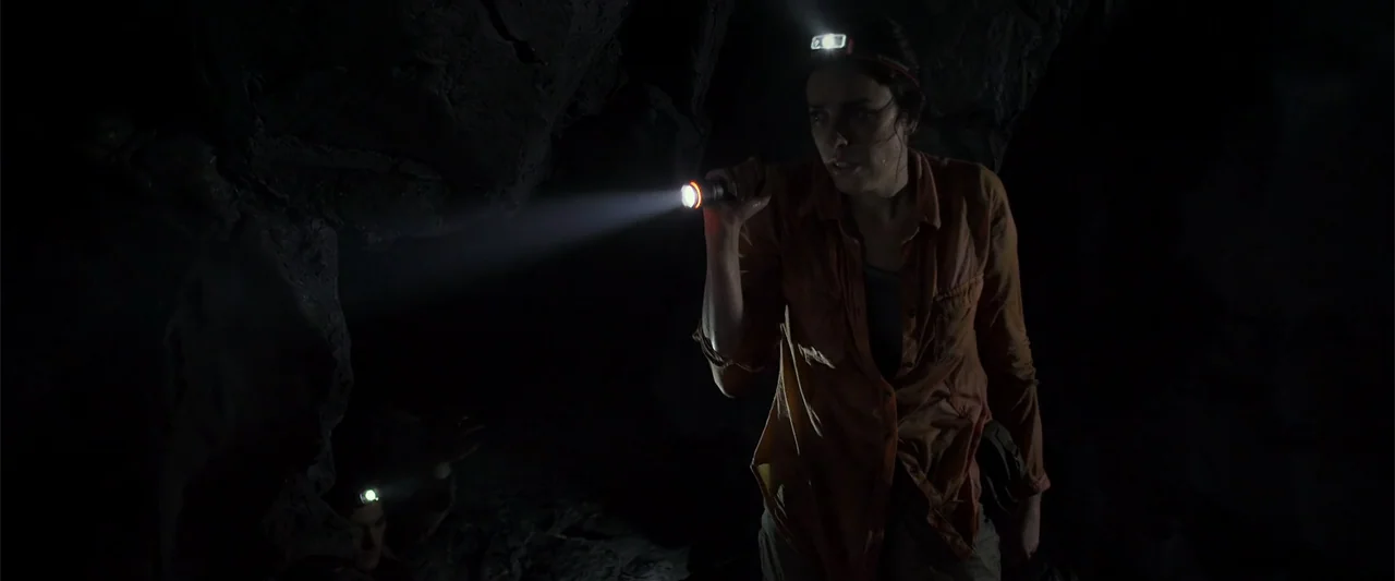 Black Water: Abyss (2020) movie still: medium — A dark, cramped stone cave interior with jagged rock walls and no natural light. A woman in an…