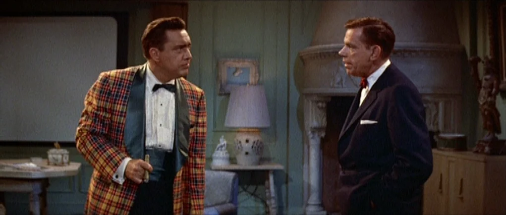 The Girl Can’t Help It (1956) movie still: medium — A mid-century living room features a stone fireplace, a table lamp, and paneled walls. Two men…