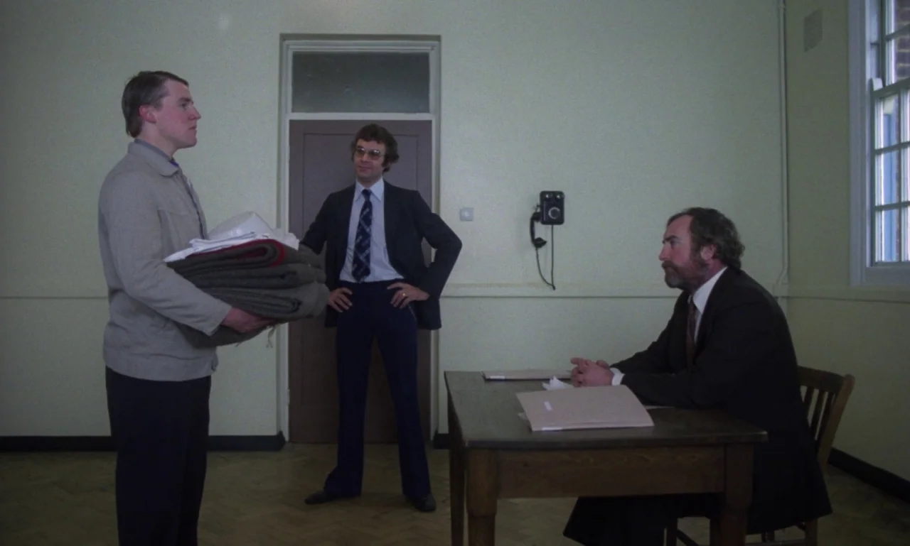 Scum (1979) movie still: fullbody — A spartan room with beige walls and a herringbone wooden floor contains a wooden desk. Three men in suits or…