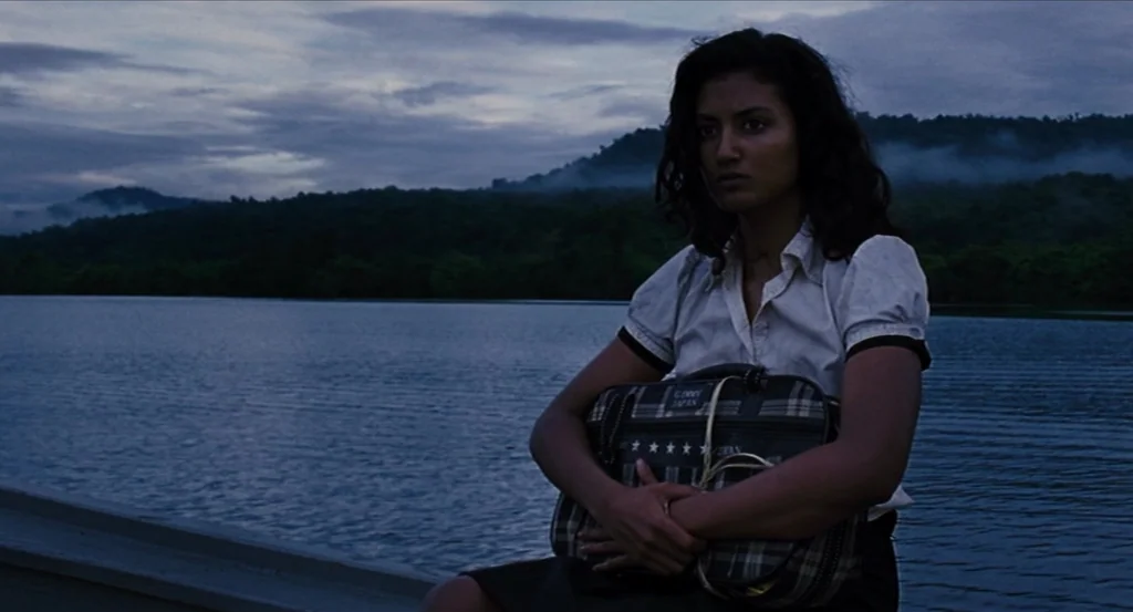 Almayer’s Folly (2011) movie still: medium — A wooden dock extends over a dark body of water surrounded by misty mountains. A woman sits on the edge…